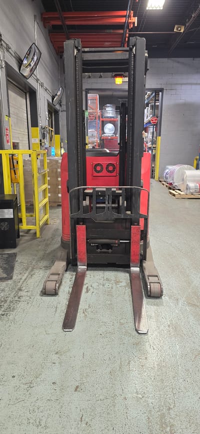 In Drummondville, Canada, a red and black reach forklift is parked in a warehouse. The forklift has two forks extended forward and two wheels visible. In the background, there are shelves stocked with various items, including large rolls wrapped in plastic. A yellow safety barrier with a fire extinguisher mounted on it is to the left of the forklift. The floor is a light gray concrete.