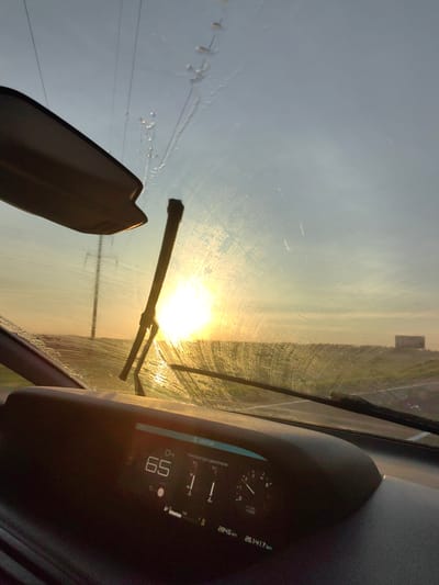 The image shows a view from inside a vehicle, looking through the windshield at a sunset. The sun is low on the horizon, casting a bright, warm glow that reflects off the wet windshield. A windshield wiper is positioned in the lower center of the frame, partially obscuring the view of the road ahead. The dashboard of the car is visible in the foreground, displaying a digital speedometer reading "65" and other information, including what appears to be trip mileage and total mileage, reading "284.5 km" and "26141.7 km" respectively. In the background, a power line tower is visible, and a building or structure can be seen in the distance on the right side of the image. The sky is clear, with some streaks of light suggesting the presence of thin clouds or atmospheric effects. The overall impression is a scenic drive during sunset, with the car's interior elements in focus in the foreground. The location is identified as Dzyarzhynsk, Belarus.