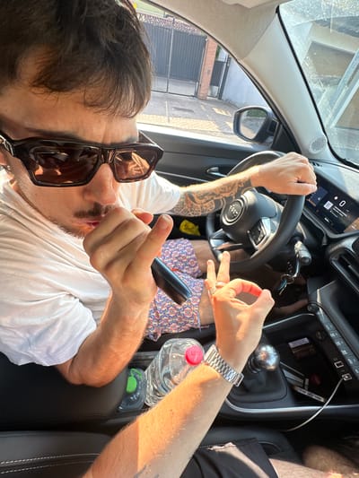 A man is in the driver's seat of a car, wearing sunglasses and a white t-shirt, with tattoos on his arm. He holds a black object to his mouth with his right hand, while his left hand gestures with two fingers up and a circle formed by his thumb and index finger. The car's interior shows a steering wheel with a digital display, a gear stick, and a water bottle. The exterior view through the window reveals a street with a fence and buildings, consistent with a residential area in Rudiano, Italy. The lighting suggests it is daytime.