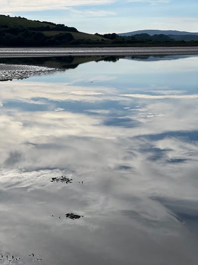 A serene scene unfolds at low tide in Dunuisce, Ireland, where the calm waters of a bay perfectly mirror a cloudy sky. The foreground is dominated by the reflective surface of the water, broken only by small, dark clumps of seaweed or debris. In the middle ground, a strip of exposed, wet sand gleams under the diffused sunlight, with patterns created by the receding tide. Beyond this, the landscape rises into rolling green hills covered in dark, dense foliage. Further in the distance, the silhouettes of larger, mist-shrouded hills form the horizon line under a pale blue sky dotted with wispy white clouds. The overall impression is one of tranquility and natural beauty, emphasizing the interplay between water, land, and sky. There are no people or animals visible in the frame.
