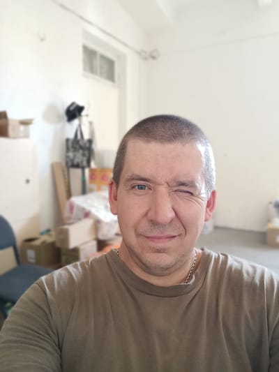 This is a close-up, selfie-style medium shot of a Caucasian man with a shaved head and light blue eyes. He is winking and smiling. He is wearing a plain, olive-green t-shirt and a thin silver chain necklace. The background is blurred and appears to be an indoor setting, possibly a storage room or a space undergoing renovation. There are several cardboard boxes of various sizes, some stacked. A blue chair is visible on the left. A bag with a patterned design hangs on a pole in the background. The lighting suggests it is daytime, and the overall mood is casual and playful due to the man's expression. There is no visible text in the image. The location context is Berdiansk, Ukraine.