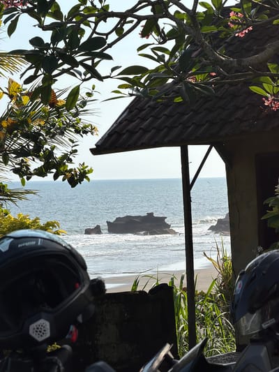 The image depicts a scenic view of the ocean from a beachfront location in Selemadeg Barat, Indonesia. The foreground is dominated by the close-up view of two motorcycle helmets, one black with white and silver detailing on the left and another black helmet with a red and white logo on the right, positioned on what appears to be a stone wall or barrier. Beyond the helmets, a sandy beach stretches towards the ocean, where moderate waves are breaking against dark, rocky outcrops in the water. The sea is a clear blue, meeting a light blue sky with no visible clouds. Partially obscuring the view is a rustic structure with a tiled roof and wooden supports, characteristic of Indonesian architecture. Lush green foliage, including palm fronds and flowering plants with pink blossoms, frames the scene from above and the sides, suggesting a tropical environment. A single bird is visible in flight over the ocean. The overall impression is one of a tranquil coastal setting, possibly a stop for travelers.