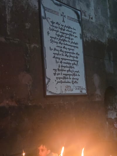 An image shows a religious plaque with Armenian script mounted on a stone wall. The plaque appears to be aged, with some signs of wear and discoloration along the edges. At the top of the plaque is an ornate design, and below it, an Armenian cross. The text on the plaque is in dark ink. Below the text, there is a decorative border and a date, "25.09.2007". The plaque is positioned on a rough, dark stone wall, suggesting an interior location such as a church or monastery. In the foreground, a child's face is partially visible, illuminated by the warm glow of nearby candles. The lighting suggests the image was taken in low-light conditions, with the candles providing the primary light source. The overall scene conveys a sense of solemnity and reverence, with the religious text and the presence of lit candles. The location is identified as Vahramaberd, Armenia.
