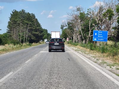 The image shows a two-lane road in a rural area, likely in or near Totleben, Bulgaria, based on the directional sign. The road surface is asphalt with white lane markings. On the right side of the road, there is a blue directional sign with white text and arrows. The top portion indicates "Бяла" (Byala) and "Русе" (Ruse) with an upward arrow, and "Russe" (Rousse) also with an upward arrow. The bottom portion shows "Тотлебен" (Totleben) with a leftward arrow. Trees line both sides of the road; the left side has a dense line of coniferous trees, while the right side has sparser deciduous trees with dry branches. In the foreground, a black SUV is driving away from the camera, straddling the white lane markings. Further ahead, a white truck is visible. The sky is blue with scattered white clouds. There are also traffic signs, including a speed limit sign of 60. The environment appears dry and sunny.