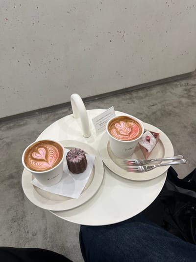 Here is a detailed description of the image:

The image shows a white, multi-tiered table with two cups of latte art and two small desserts. Each cup features a pink-colored latte art design. One tier of the table has a piece of pink cake topped with a white substance and what appears to be a preserved cherry blossom, accompanied by two forks. The other tier has what looks like a canelé.  

The table is placed on a concrete floor, and a person in denim jeans and a black jacket is seated next to it. A small card, possibly a menu or receipt, is visible near one of the cups. Part of the text on the card is legible: "butter".

There is no information about the location being Nakano, Japan in the image.