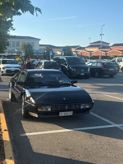 The image captures a parking lot scene in Serravalle Scrivia, Italy, under a clear blue sky, suggesting it is daytime with good weather. The primary focus is a line of parked cars, including a dark-colored vintage Ferrari in the foreground, identifiable by its distinctive grille and headlights, and the license plate "AY 353 YC". Other vehicles visible include SUVs and vans, one of which has a license plate starting with "DC 348FA". In the background, there are buildings with terracotta-tiled roofs and modern architecture, hinting at a commercial or shopping area. The presence of streetlights and road markings, including parking lines, confirms the setting as a public parking lot. A few individuals can be seen, some standing near the vehicles, but their activities are not clearly defined. The overall impression is a mundane scene of cars parked in a sunny outdoor environment.