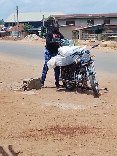 In Iseyin, Nigeria, a man is standing beside a motorcycle loaded with goods. He is wearing a black hooded sweatshirt with a graphic design on it, blue patterned pants, and sandals. The motorcycle is blue and white and has a license plate that reads "EDUCATOR". It is carrying several large bags, one of which has "WHEAT FLOUR" printed on it, as well as some boxes. The scene appears to be outdoors on a dirt road, with buildings and trees visible in the background. The sky is overcast, suggesting it might be a cloudy day. The man's posture and the way he is looking at the goods might indicate he is preparing to transport them or has just arrived with them. The overall impression is of a busy day with commercial activity.