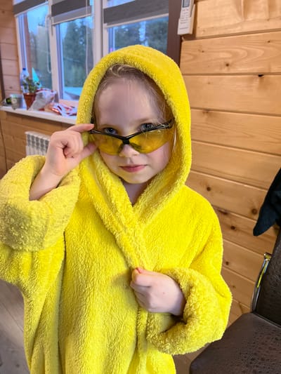 A child with fair skin and blonde hair wears a yellow hooded bathrobe and yellow-tinted sunglasses. The child is holding the sunglasses frame with one hand and resting the other hand on the bathrobe. The background consists of light brown wooden paneling. The photograph appears to be taken indoors. The location, Bol'shakino, Russia, is not evident in the image.