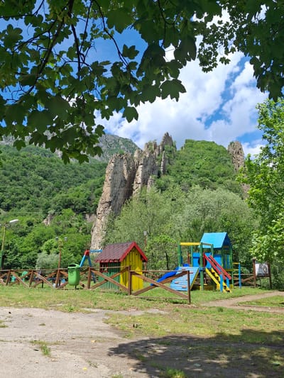 This image captures a vibrant outdoor scene in Lyutibrod, Bulgaria, showcasing a blend of natural beauty and a recreational area. The perspective is looking slightly upwards, with lush green tree leaves framing the top and left sides, partially obscuring the bright blue sky.
The background is dominated by dramatic, rugged rock formations and cliffs, characteristic of a mountainous or hilly region. These rocky outcrops are interspersed with and surrounded by dense, verdant deciduous forest that covers the sloping terrain. Above, the sky is a clear blue with scattered white, fluffy clouds, indicating pleasant weather.
On the right side of the image, a colorful children's playground is prominently featured. It consists of multiple wooden structures painted in vivid primary colors: bright blues for slides and platforms, yellows and reds for climbing stairs, and green accents. There are several small house-like play structures integrated into the design. A sturdy wooden fence separates the playground from a light-colored dirt path that runs along the right side of the frame, lined with grass. A green trash can, adorned with a white circular symbol (likely a recycling or disposal logo), is attached to this fence. In the distance beyond the playground, there are hints of utility poles or lighting fixtures, and a small, unreadable sign.
No people are visible in the scene, suggesting a quiet moment. The bright sunlight casts shadows on the path, indicating it is daytime, likely mid-day. The overall impression is one of a peaceful, inviting outdoor space, perfect for recreation amidst a striking natural landscape. No legible text is visible in the image.