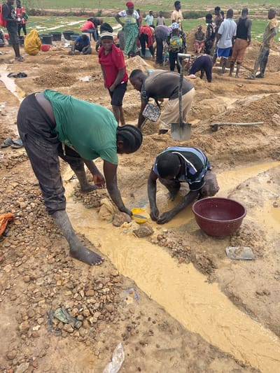 The image shows a group of people engaged in artisanal mining activities in Maiyanga, Nigeria. The scene is a muddy, rocky area with a shallow stream of dirty water flowing through it. Several individuals are working along the water's edge, some digging, others sifting through the sediment, and one using a yellow scoop. There is a large, dark-colored bucket near one of the miners. Most people are barefoot or have mud-covered feet and legs. The individuals are wearing casual clothing. In the background, there is a larger group of people also involved in the mining operation, and some greenery or vegetation is visible in the far distance. There are several dark-colored buckets in the background as well. A shovel is visible nearby.