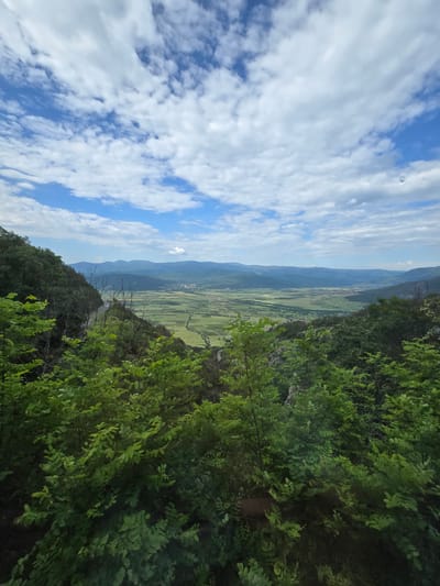 A high-angle, panoramic view captures a vast rural valley landscape near Karnare, Bulgaria. The upper portion of the image is occupied by a partly cloudy sky, featuring clear blue sections interspersed with numerous white cumulus clouds. The immediate foreground is dominated by dense, vibrant green foliage from trees and shrubs. Below this vegetation, a wide, fertile valley unfolds, characterized by extensive agricultural fields arranged in geometric patterns of varying green shades. A small village or settlement, identifiable by its clustered buildings with reddish-brown rooftops, is visible in the mid-distance within the valley. The background features a range of mountains, appearing in muted blue and grey tones due to atmospheric haze, forming the distant horizon. No human subjects or specific actions are depicted within the scene.