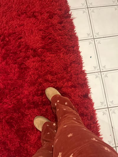 A high-angle shot shows a person standing on a vibrant red shag rug. The rug extends from the lower left edge, covering most of the frame. The person wears beige slip-on shoes and reddish-brown pants with a floral pattern. The upper left corner features a section of white tiled floor with a decorative gray pattern at the corners of each tile. The perspective is slightly tilted, giving a dynamic feel to the composition.