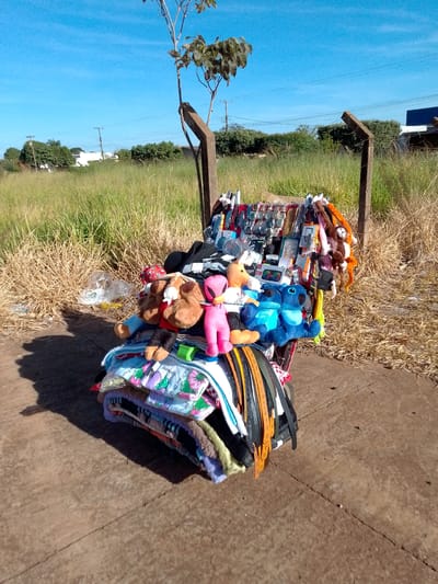 An outdoor, eye-level, wide shot captures a makeshift vendor stall situated on a concrete surface near a grassy field under a clear blue sky. The stall, constructed from scavenged materials including two wooden posts and a metal frame, is laden with a variety of merchandise. Prominently displayed are numerous stuffed animals, including a pink one, a brown bear, a blue Stitch character, and a monkey. Alongside these toys are an assortment of accessories such as sunglasses, watches, and what appear to be small electronic devices, all arranged on shelves and hanging from the structure. Beneath the displayed items, a pile of folded fabrics, including what looks like blankets or clothing with patterned designs, is visible. The background reveals a dry, grassy field with some scattered trash, followed by a line of trees and bushes. In the distance, buildings and utility poles are visible, hinting at an urban or suburban setting within São Gabriel Do Oeste, Brazil. The scene suggests a casual, open-air retail operation.