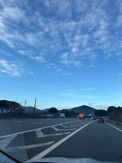 A highway scene under a blue sky with wispy clouds. A mountain rises in the background, alongside the road lined with trees and power lines. A vehicle drives along the highway, as seen from the passenger seat of another car. Road markings, including dashed lines and chevron patterns, are visible on the asphalt. The sky takes up a significant portion of the frame.