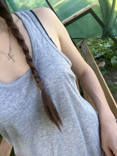 The image shows a close-up, upper-body shot of a person wearing a grey tank top and a silver cross necklace. Their brown hair is styled in a braid that hangs over their shoulder. The person appears to be sitting on a wooden chair outdoors, with a green mesh structure and foliage visible in the background. The scene is set in the city of Postoyata, Ukraine.