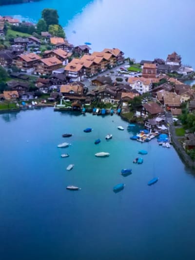The image shows an aerial view of a lakeside town with traditional European-style architecture. The town features buildings with red-tiled roofs, many of which are clustered along the hillside overlooking a body of water. The water is a vibrant blue and is dotted with numerous boats, many of which are covered with blue tarps. Some boats are moored near docks and jetties that extend into the water. The surrounding landscape includes green hills and trees. There are no people visible in the image. The style of architecture and the presence of a lake suggest a European setting, possibly in the Alps or a similar mountainous region. This location is not in Edmonton, Canada.