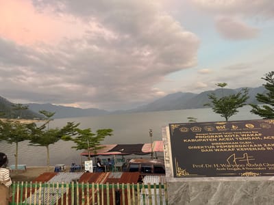 The image depicts a serene lakeside scene in Mendale, Indonesia, with a focus on a black plaque commemorating the "PROGRAM KOTA WAKAF KABUPATEN ACEH TENGAH." The plaque, adorned with gold accents and official emblems, prominently displays the inauguration date as November 8, 2020, and names Prof. Dr. H. Waryono Abdul Ghafur as the officiant.
In the foreground, a person with dark hair, wearing a striped shirt and a brown apron, stands facing away from the camera, looking towards the lake. Behind them, a rustic fence made of bamboo and corrugated metal separates the viewing area from the lakefront. Several small trees with manicured foliage are planted along the shore, adding a touch of green to the landscape.
The lake itself occupies a significant portion of the image, with calm waters reflecting the overcast sky and distant mountains. The sky is filled with dramatic, wispy clouds, some tinged with a soft pink hue, suggesting either sunrise or sunset. Several small covered stalls and tables with chairs are set up along the water's edge, indicating a recreational or dining area. A rusty brown van is parked near one of these stalls.
The background is dominated by a range of majestic, undulating mountains that recede into the distance, their peaks softened by atmospheric haze. The overall impression is one of natural beauty and a quiet, communal gathering place. The presence of the plaque suggests a significant event related to Islamic endowment (wakaf) and community development has taken place in this location.