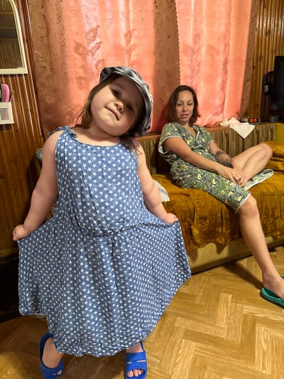 Two people are lying on a patterned couch and a peach-colored bed. A young child in a blue dress with white stars and a grey and white checkered hat is in the foreground. The child is looking up and smiling. Behind the child, a woman with dark hair is lying on the couch, wearing a green and yellow patterned outfit. Her head is tilted and she appears to be looking at the camera. The couch is a textured gold color, and the bed has a floral pattern. The floor is made of wooden planks. Blue flip-flops are visible on the feet of the child and another person off-camera to the right. The scene appears to be indoors, possibly a living room or bedroom. No text is visible in the image. The time of day and weather are not discernible. The overall emotion conveyed is relaxed and playful.