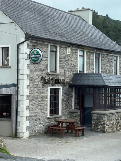 The image depicts the exterior of a stone building in Gortahork, Ireland. The building appears to be a pub or restaurant, with a wooden sign displaying "TAEG SHILL" in gold lettering. A prominent Heineken beer advertisement is also visible. The architecture features a grey slate roof and rough-hewn stone walls. White decorative crenellations run along the lower section of the building. A wooden picnic table and benches are set up outside, suggesting an outdoor seating area. The scene is set against a backdrop of lush green trees and an overcast sky, hinting at a cool, possibly damp day. The image is taken from a slightly elevated perspective, looking towards the building. There are no people visible in the frame. The overall impression is one of a traditional establishment in a rural Irish setting.
