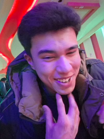 The image depicts an indoor scene in Takhtakuprik, Uzbekistan, focusing on a young adult male. His face, illuminated with a prominent purple hue, is captured in a close-up. He has dark, voluminous hair and a smiling expression, revealing his upper teeth, with his right eye (viewer's left) partially closed. He is wearing a dark, possibly padded jacket, with a visible light tan inner lining around the collar. His left hand, fingers splayed, is positioned near his chin and neck. The background is blurred, displaying abstract forms and streaks of red and green light, consistent with an entertainment venue featuring artificial colored illumination.