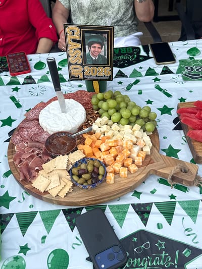The image shows a wooden charcuterie board laden with a variety of food items, including grapes, cubed cheeses, a wheel of brie with a spoon in an accompanying dish of chutney, olives, crackers, nuts, and various cured meats like salami and prosciutto. The charcuterie board is set on a table covered with a white plastic tablecloth decorated with green graduation-themed designs such as diplomas, stars, and "congrats" messages. A photo frame displaying a graduation photo and the words "Class of 2026" is visible near the board. There are two smartphones on the table, one black and one red, and what appears to be another phone or electronic device in the upper left corner. A hand wearing a red shirt is also visible.
Given the graduation-themed decorations, it is likely that this scene depicts a graduation celebration or party. Without additional context from the image, the location in Southlake, United States of America, cannot be verified.