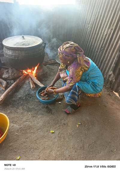 A woman with a purple and gold headscarf and a blue dress kneels beside a fire. A large black pot sits over the flames, generating smoke. She holds a blue bowl with food inside, possibly washing it. There is a yellow bucket on the ground to the left. The background is a corrugated metal wall. The scene appears to be an outdoor cooking setup.