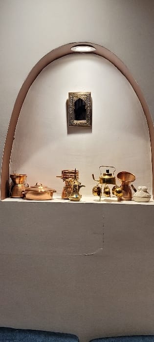 An arched niche in a light-colored wall displays several golden-toned objects, possibly brass or copper cookware, stacked vertically along the right side. A rectangular framed object, seemingly a mirror, is positioned on the left wall of the niche. A small, circular light fixture is near the top left of the arch. The wall surrounding the niche appears to be a muted taupe color. Parts of blue upholstered furniture are partially visible in the right edge of the image.