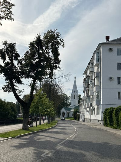 In Voksherino, Russia, an overcast day presents a view of a street lined with a tall, white apartment building on the right and a tree-lined sidewalk on the left. A white church with a tall, pointed steeple and a cross on top stands in the middle ground, partially obscured by trees. A car drives away from the viewer on the asphalt road, which has white lane markings. Several people are visible on the sidewalk, including some sitting on a park bench under a large, leafy tree on the left. The apartment building features multiple balconies and air conditioning units. Lush green foliage covers a section of the building's base on the right. The sky is filled with scattered clouds.