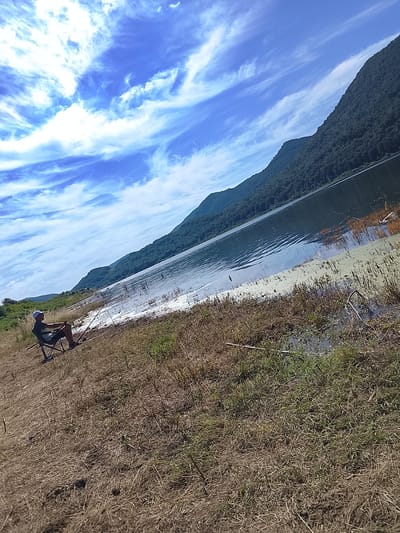 The image depicts a serene scene by a lake, likely in Asparuhovo, Bulgaria. The main subject is a person sitting in a folding chair on the grassy bank, holding a fishing rod, engrossed in the activity of fishing. The person is wearing a blue cap, a dark t-shirt, and shorts. A fishing rod is extended towards the water, suggesting an ongoing attempt to catch fish.
The setting is a natural landscape with a vast lake reflecting the sky and the surrounding forested mountains. The mountains, covered in dense green foliage, rise majestically on the far side of the lake, creating a dramatic backdrop. The sky is a vibrant blue, adorned with wispy, white clouds that streak across it, adding a sense of movement and depth. The weather appears to be fair and sunny, typical of a pleasant day. The time of day is likely late morning or early afternoon, judging by the bright sunlight.
The foreground is dominated by dry, yellowish grass and some scattered green patches, indicating a semi-arid environment or late summer season. The lake's edge shows some aquatic vegetation and possibly some debris. The overall mood is peaceful and tranquil, evoking a sense of calm and solitude. The gentle ripples on the water's surface suggest a light breeze. There is no visible text in the image.