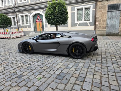 A grey Lamborghini Aventador is driving on a cobblestone street in front of a building with a stone facade and a grey-and-white patterned wall. The building has arched doorways and windows with ornate grilles. A round, green tree stands to the right of the car, and a temporary construction barrier with orange and white stripes is visible on the left. A traffic sign depicting a shovel and a pile of dirt is positioned near the building's entrance. The car's driver, a man with a light-colored shirt and sunglasses, is visible through the window. The scene appears to be in a historic district, likely in Ansbach, Germany, given the architectural style.