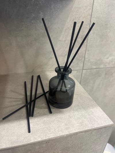 The image displays a gray glass reed diffuser bottle filled with a dark liquid, placed on a light gray tiled surface. Several black reed sticks are inserted into the bottle, with some also scattered on the surface in front of it. The background consists of a light gray tiled wall. The scene is indoors, likely in a bathroom or a room with similar decor. There are no people or animals present. The lighting appears to be artificial, creating subtle shadows. The image does not provide any information that would allow for identification of its location within the city of Староживотинное, Russia.