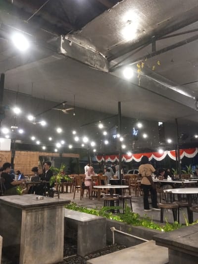 This image depicts an evening scene within a partially open-air cafe or restaurant in Pontianak, Indonesia. The overhead structure features a dark, flat ceiling with exposed metal ventilation ducts and numerous string lights providing illumination. Multiple ceiling fans are suspended from the ceiling. The floor appears to be a mix of concrete and gravel, with concrete planters containing green foliage in the foreground.
Approximately 15 to 20 individuals are present, seated at various wooden tables with white tabletops. The clientele consists of both men and women, some wearing headscarves (hijabs). Individuals are engaged in social interaction, dining, and using personal electronic devices such as laptops and mobile phones.
In the mid-ground, a female staff member, identifiable by a light-colored uniform shirt and a dark apron, stands facing away from the camera, observing the dining area.
The background includes a wooden slatted wall and, further back, a section adorned with decorative red and white drapes, possibly indicating a celebration or national theme. The overall environment suggests a casual, bustling atmosphere.