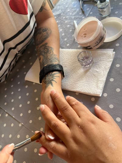 The image shows a close-up, overhead view of a person's hands being worked on, presumably for a nail treatment. One arm, adorned with tattoos, is visible on the left side of the frame, wearing a black bracelet. The other arm, with bare nails, is positioned in the center and right, being manipulated by the first person's hands. A nail brush with a small amount of acrylic product is being applied to one of the nails.
Scattered on a grey, polka-dotted surface are various nail treatment supplies: a jar of light pink acrylic powder labeled "NEXUS MAKE UP TO NATURAL", a small clear glass dish, a white paper towel, a bottle of nail primer or dehydrator, and a pair of nail clippers.
The scene suggests a personal nail care or salon setting. No specific location in Figueres, Spain can be determined from the provided image.