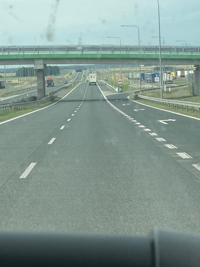 The image depicts a highway scene in Zabiele, Poland, with a focus on the road and its surroundings. The foreground shows the asphalt of the highway, marked with white dashed lines and solid white lines. In the middle ground, several vehicles are visible, including a red truck and a white camper van driving on the highway. An overpass, painted green and grey, spans across the highway in the background. On the right side of the highway, a rest stop or parking area is visible, with several trucks parked. The sky is overcast. The image appears to be taken from inside a vehicle, looking forward.