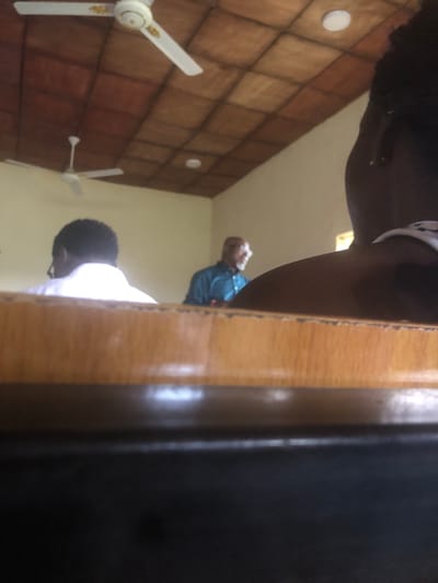 In a dimly lit room in Laminga, Nigeria, a person's head and torso are visible in the foreground, obscured by the camera's perspective. The person appears to be wearing a white garment with black polka dots. In the background, two ceiling fans are visible, one closer and larger than the other. The walls are covered in a tiled pattern of warm brown and beige squares. In the middle ground, two individuals are partially visible. One, wearing a blue shirt, is looking upwards and away from the camera. Below them, another person wearing a white shirt is also looking downwards and away. The overall scene suggests an indoor environment, possibly a room in a residential or communal building. The lighting is soft and ambient, with no strong indications of time of day or weather conditions. The image is angled, as if taken from a low vantage point looking upwards.