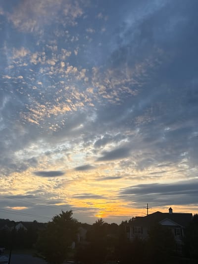 A dawn sky glows with soft oranges and yellows near the horizon, transitioning to a muted blue overhead. The sky is populated with wispy, scattered clouds, interspersed with darker, more solid cloud formations. Silhouetted against the vibrant sky are the dark shapes of trees and the rooflines of houses. One house, partially visible in the lower right quadrant, has a distinctive peaked roof and is surrounded by trees. Utility lines are visible stretching across the frame. The scene depicts a suburban landscape at sunrise in Allen Township, United States of America.