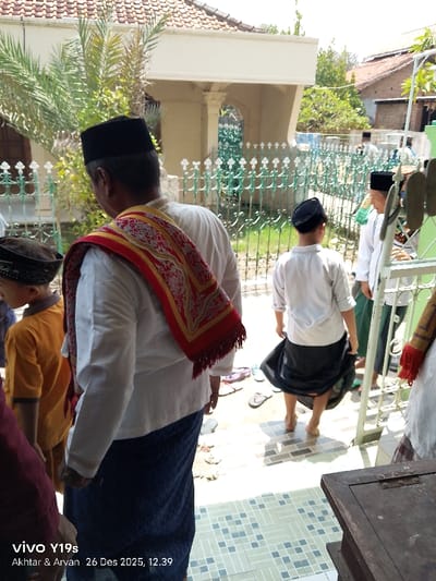 A group of Muslim men and boys are gathered, presumably for prayer. They wear traditional attire, including sarongs, white shirts, and peci hats. A man in the foreground has a red and gold shawl draped over his shoulder. Most have removed their shoes, leaving them scattered on the tiled ground. A fence and building can be seen in the background. A timestamp indicates the photo was taken on December 26, 2025.