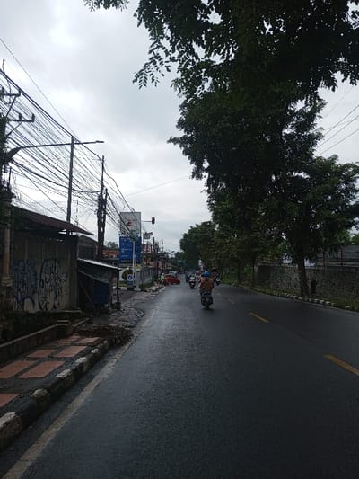 The image shows an urban street scene in Kamji Na-Yekha, Bhutan, under an overcast sky. The asphalt road surface appears wet, suggesting recent rain. A single individual on a light-colored scooter, wearing a beige helmet and light jacket, is riding away from the camera in the right lane of the two-lane road, which is marked with a broken yellow line on the right and a solid white line on the left. Several other motorcycles are visible further down the road, also moving away from the viewer.
On the left side of the street, a raised concrete sidewalk with red and black square tiles is partially covered in dirt. Behind the sidewalk, a grey concrete wall is extensively covered with blue and white graffiti. Above this wall, a building with a corrugated metal roof is partially visible. A tall utility pole stands on this side, densely clustered with numerous black electrical and communication cables, and features a street light. Further along the street, a commercial building with a prominent blue and white sign displaying white text is visible, located near a red traffic light. A red car is also present near these structures.
The right side of the road is bordered by a continuous line of mature trees with dense green foliage, which rise above a stone wall running along the roadside. The overall environment suggests an active urban area.