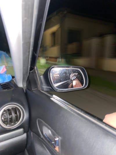 This is a nighttime, motion-blurred photograph taken from inside a vehicle, likely in the city of Yasen, Bulgaria. The camera is positioned to capture the interior of the car and a reflection in the side-view mirror.
In the foreground, the dark, textured interior of the car door is visible, including a circular air vent with metallic slats and a chrome door handle. To the left, a bright, silver-colored pillar of the car's frame is prominent. A portion of a blue stuffed toy with pink accents is visible on the dashboard or near the windshield on the left side.
The side-view mirror, positioned on the right side of the image, reflects a blurry image of a person's hand and arm. The hand appears to be making a peace sign, with two fingers extended. The reflection also shows the interior of the car and possibly the person's leg or footwear.
The background, seen through the car window and in the side-view mirror, is heavily motion-blurred, indicating the vehicle is in motion. It depicts a dimly lit urban or suburban environment, with the blurred shapes of buildings and possibly streetlights. The overall impression is that of a passenger capturing a moment while traveling at night.