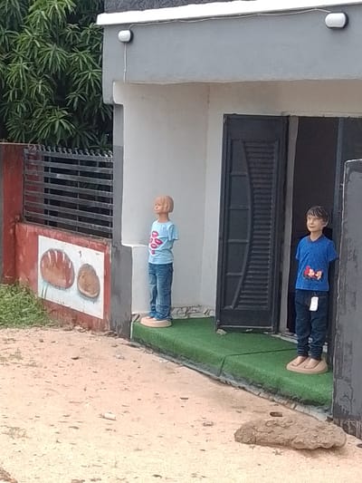 The image depicts a storefront in Gulum, Nigeria. Two child-sized mannequins are positioned outside the entrance. The mannequin on the left is wearing a light blue t-shirt with a pattern of red lips and blue jeans. The mannequin on the right is dressed in a blue t-shirt with a graphic of a mouse and dark blue jeans. Both mannequins stand on beige oval bases, with the right mannequin standing on a green artificial turf mat that extends from the storefront. The building has a grey facade with a dark-colored, slatted door. To the left of the entrance, there is a red-painted wall with a mural of two loaves of bread. A metal gate with horizontal bars is visible behind the left mannequin. Lush green foliage from a tree is seen to the left of the gate, suggesting an outdoor setting, likely during the day, with overcast weather. The ground in front of the store is sandy and uneven, with a few scattered pebbles and a larger rock in the foreground. No discernible text is visible in the image.