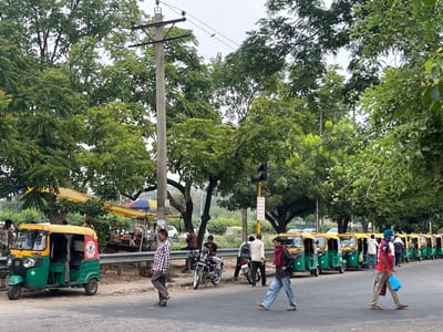The image depicts a street scene in Kajheri, India, with a prominent row of green and yellow auto-rickshaws parked along the roadside. Several people are visible, some walking across the street, others standing near the auto-rickshaws or under roadside awnings. A traffic light is present, as are utility poles and overhead wires. Lush green trees line the street, suggesting a semi-urban or suburban environment. The sky is overcast.
