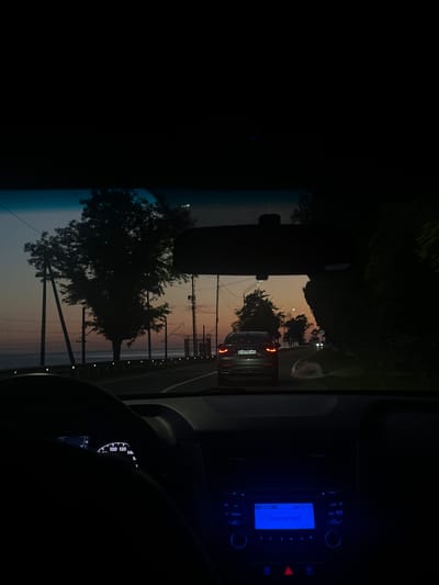 This media file captures a view from inside a vehicle, looking out the windshield at dusk. The scene depicts a road with cars ahead, silhouetted against a soft, pastel-colored sky. Trees line the side of the road, their branches also dark against the fading light. Power lines and poles are visible, suggesting an urban or suburban setting.

A dark-colored car with its brake lights illuminated is in the foreground, traveling in the same direction. Further ahead, another vehicle's headlights are visible. The interior of the car includes the dashboard with a radio displaying "Connected" in blue text, and a partially visible speedometer. The steering wheel is also visible, indicating the perspective of the driver or a passenger in the driver's seat.

The time of day is clearly evening, with the sky exhibiting hues of orange, pink, and light blue, characteristic of sunset or twilight. The overall mood is tranquil, perhaps even serene, due to the soft lighting and the gentle progression of the drive. The location is contextually noted as Primorskoe, Abkhazia.