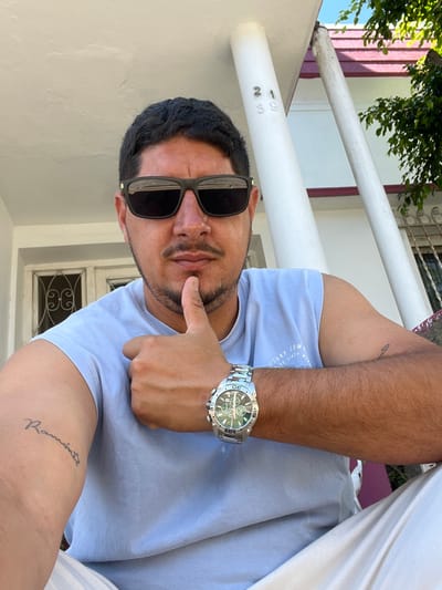 A male subject is depicted from the chest up, seated outdoors in Bernal Este, Argentina. He has dark hair, a mustache, and stubble, and wears black rectangular sunglasses. He is dressed in a light blue sleeveless t-shirt with a faint graphic design. A silver-toned wristwatch with a prominent bezel and multiple sub-dials is worn on his left wrist. A script tattoo spelling "Ramón" is visible on his left bicep. His left hand is raised, with the thumb extended and resting against his chin. Light-colored pants are partially visible at the bottom of the frame. The background features a white architectural column displaying the numbers "21" and "39" vertically. Further back, parts of a building are visible, including a white window with a decorative metal grate, a reddish-brown roofline, and green foliage. The scene is illuminated by bright natural daylight.