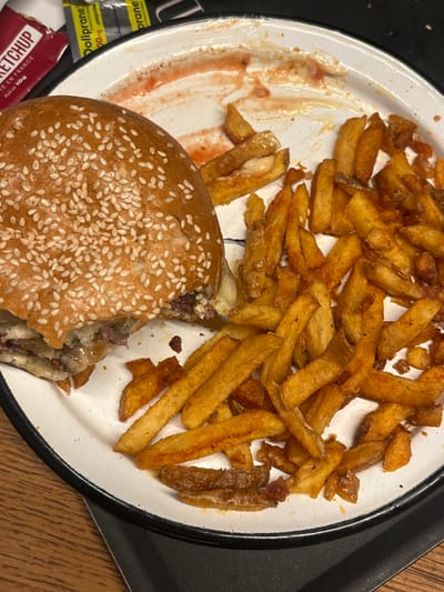 The image displays a partially consumed meal on a white plate with a black rim, situated on a dark surface that rests upon a wooden table. The primary subjects are a large, partially eaten burger and a serving of French fries. The burger, positioned on the left side of the plate, features a sesame seed bun, melted cheese, and visible meat patties; a bite has been taken from its left side. To the right and surrounding the burger are numerous golden-brown French fries, many appearing seasoned. Red sauce residue, consistent with ketchup, is smeared on the upper right portion of the plate. In the upper background, a red ketchup sachet labeled "KETCHUP" and "FABRIQUÉ EN FRANCE" is visible. Adjacent to it are two silver and blue sachets, one identifiable as "Doliprane paracétamol 1000 mg." No individuals or active interactions are depicted. This scene is located in Serris, France.