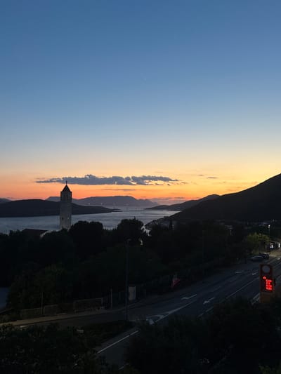 The image depicts a sunset over the Adriatic Sea, likely taken from a higher elevation point in Neum, Bosnia and Herzegovina. To the left, a blue sky fades into orange and yellow hues near the horizon. A line of clouds stretches vertically through the center of the frame, reflecting the sunset colors.

A mountain range runs parallel to the coastline on the right. The sea is visible in the middle, reflecting the sky. A white church tower with a cross at the top stands prominently against the background of the water and mountains.

A road runs along the coastline on the right, with trees lining both sides. A gas price sign is visible on the right side of the road.
