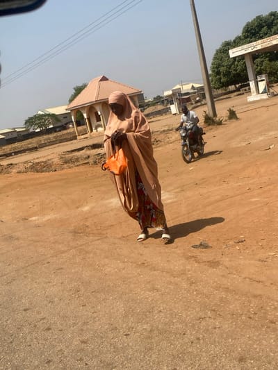 This image, captured in Lapai, Nigeria, depicts an outdoor street scene under clear skies and strong sunlight. In the foreground, a woman, clad in a light beige full-length garment covering her head and body, with a colorful patterned dress visible underneath, stands on a reddish-brown dirt road. She wears white open-toed sandals, holds an orange bag with black straps in her left hand, and positions her right hand near her chest, with her gaze directed downwards. In the mid-ground, a man rides a dark-colored motorcycle, moving from right to left across the frame; he wears a white t-shirt and dark trousers. The background features a small, light-colored building with a pyramidal roof and arched openings on the left, and a white gas station canopy with a partially legible sign reading "PETROL... RESOURCES" and a fuel pump on the right. A tall grey utility pole stands in front of the gas station, and sparse trees and additional low-rise buildings are visible in the distant background. Distinct shadows are cast on the ground by the strong overhead sun.
