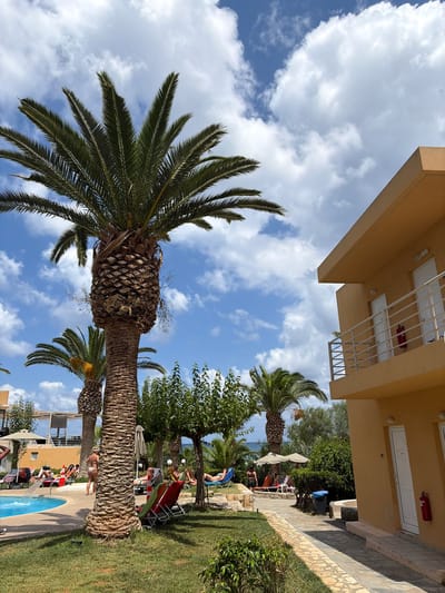 The image shows a sunny day at a resort in Vlichada, Greece. The foreground is dominated by a large palm tree with a textured trunk. To the right, a two-story building with a tan facade and balconies with white railings is visible. The building has several white doors, two of which are clearly numbered (417 and a faded number above another door). A red fire extinguisher is mounted on the wall near one of the doors. Beyond the building, a swimming pool with clear blue water is partially visible, with lounge chairs and umbrellas set up around it. Several people are seen relaxing on lounge chairs by the pool and on the grassy area. Palm trees and green foliage are scattered throughout the scene, with the blue sky and white clouds providing a backdrop. In the distance, the sea can be seen. The overall atmosphere is one of leisure and vacation.