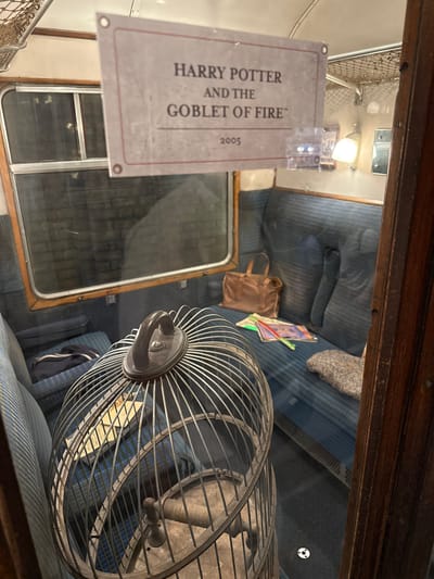 This media file shows the interior of a train carriage, possibly a prop from a Harry Potter exhibition given the sign visible. The carriage has blue striped seats and appears to be set up as a display.
**Subjects:**
* **Objects:** A metal fan is prominently displayed in the foreground, angled towards the viewer. On the seat behind it are several items, including a brown bag, some books or magazines, and a grey, fuzzy object (possibly a blanket or cushion). A sign is affixed to the window on the left.
* **Scene/Setting:** The scene is the inside of a vintage train carriage. The seats are upholstered in a blue fabric with darker blue stripes. The walls and ceiling are visible, with overhead lighting fixtures. A window is present on the left, offering a view into the carriage. The overall impression is of a preserved or recreated historical environment.
**Activity or Event:**
There is no apparent activity or event taking place; the scene appears to be staged as a static exhibit.
**Notable Details:**
* **Location Cues:** The sign mentions "HARRY POTTER AND THE GOBLET OF FIRE" and the year "2005," strongly suggesting a connection to the Harry Potter franchise and likely an exhibition or film set. The interior styling of the train carriage is reminiscent of older British trains.
* **Lighting:** The interior lights are on, creating a well-lit but slightly diffuse atmosphere.
* **Emotional Tone:** The image conveys a sense of nostalgia and perhaps a touch of mystery due to the cinematic association.
**Visible Text:**
The most prominent text is on the sign, which reads:
"HARRY POTTER
AND THE
GOBLET OF FIRE™
2005"