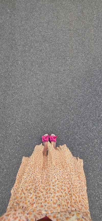 A top-down perspective of a person's feet and lower legs wearing a flowing, light-colored, floral-patterned maxi dress and bright pink bow sandals. The individual is standing on an asphalt surface. The image provides no information about the city of Lieli, Switzerland.