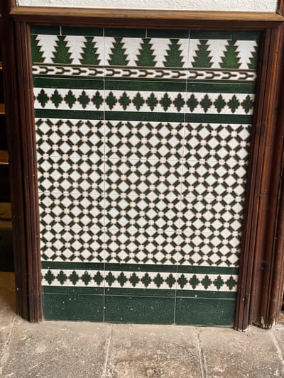 An eye-level shot captures a decorative ceramic tile panel framed by dark brown wooden molding, likely part of an interior or sheltered exterior wall in San Cristóbal De La Laguna, Spain. The tile panel displays a symmetrical, intricate design primarily in dark green and white.
The central and widest section features a repeating geometric pattern of dark green squares rotated 45 degrees, interconnected by smaller white squares and star-like shapes, creating a lattice effect. Flanking this central pattern are two narrower vertical bands, each showcasing a series of dark green trefoil or quatrefoil-like shapes outlined in white, set against a white background. The outermost vertical bands (closest to the solid dark green edges) are composed of a repeating zigzag or stepped pattern in dark green against white, resembling stylized trees or arrows. The entire tile arrangement is subtly outlined in a lighter, off-white or beige tone.
To the left of the wooden frame, a plain white wall is visible. To the right, a rough-textured, light gray or beige stone wall or pillar suggests an older architectural structure. The tiles exhibit a slightly glossy finish with minor imperfections, while the wooden frame shows its natural grain. The lighting is soft and even. No people or visible text are present in the image.